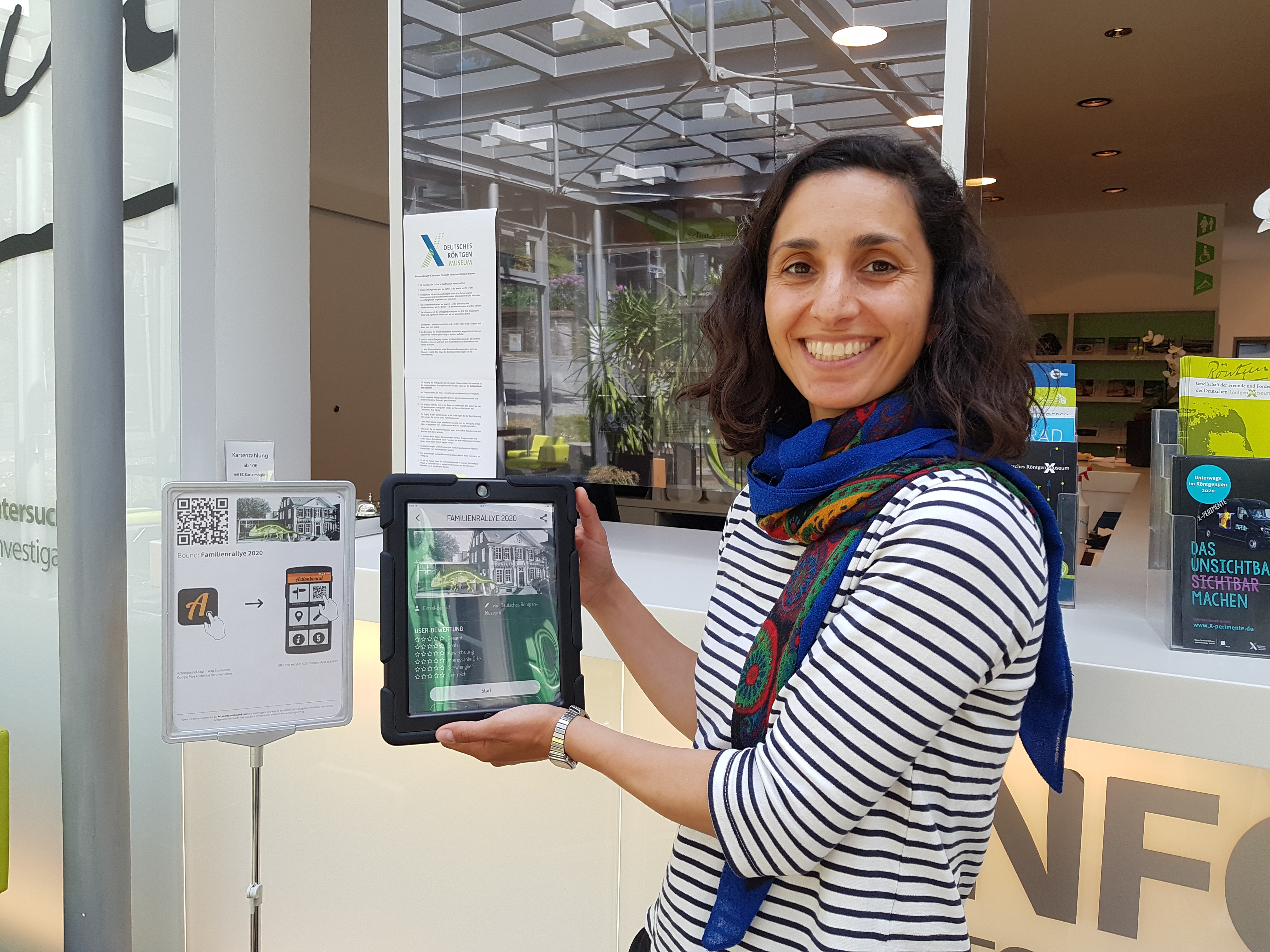 Einführung actionbound Frau mit Tablet in der Hand im Foyer des Röntgen Museums im Hintergrund ein Bild mit dem Actionbound Logo